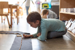Boy in blue shirt kneels on wooden floor and reaches out to touch a string of gold beads on a gray rug.
