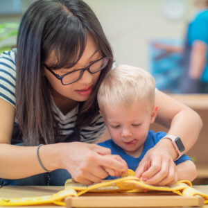 A woman with dark hair and a blue and white striped shirt leans over to help a small boy as he learns to button