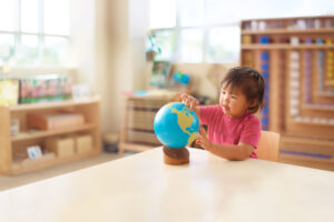 A young girl with black hair and a pink t-shirt sits at a light wood desk and points to a globe