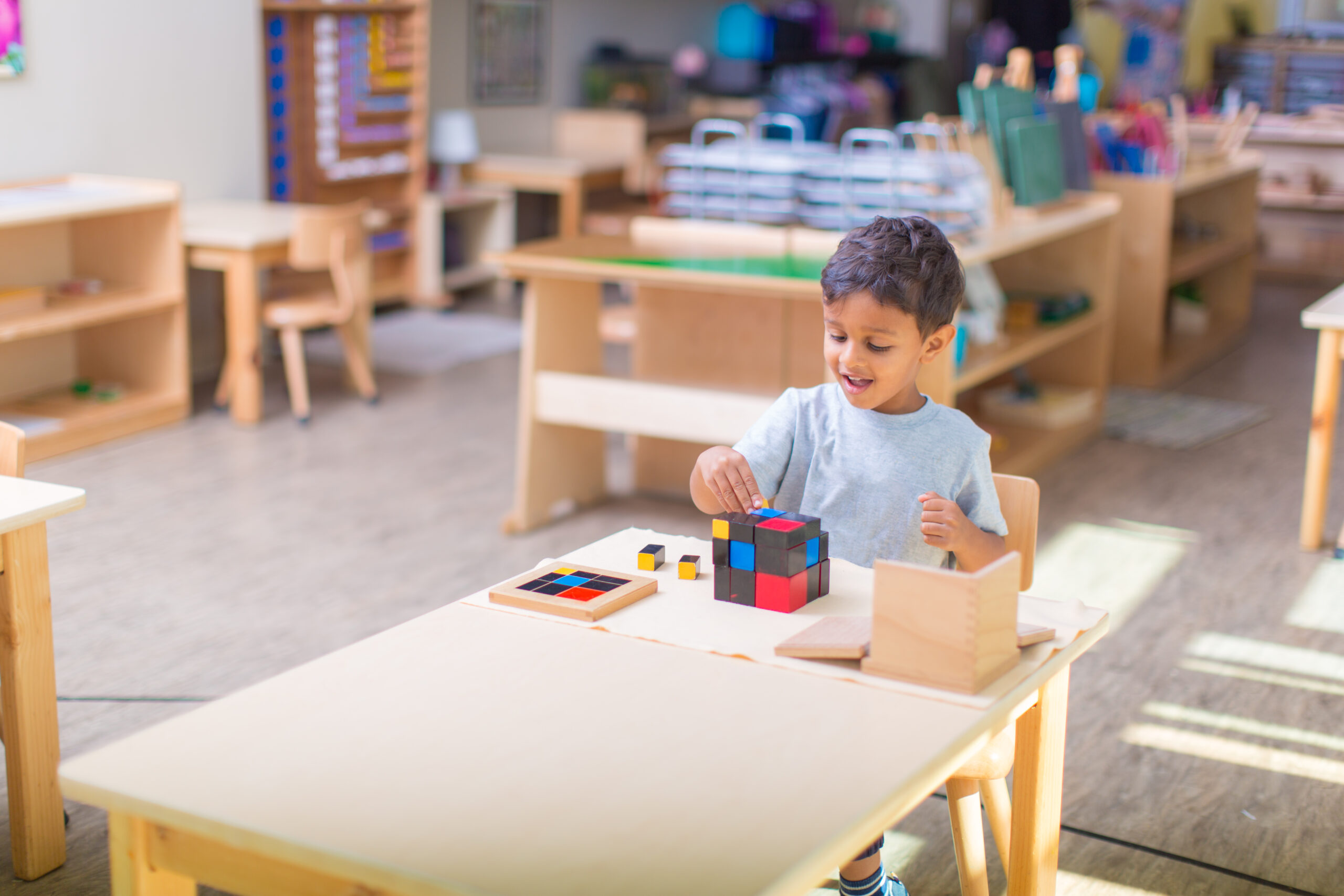 Young boy with grey t-shirt sits at wooden desk and works with a trinomial cube that is colored blue, black, and red