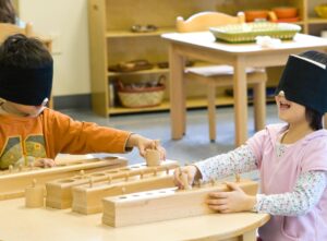 Two young children wear blindfolds and try to put round cylinders into the correct hole in a wooden box.