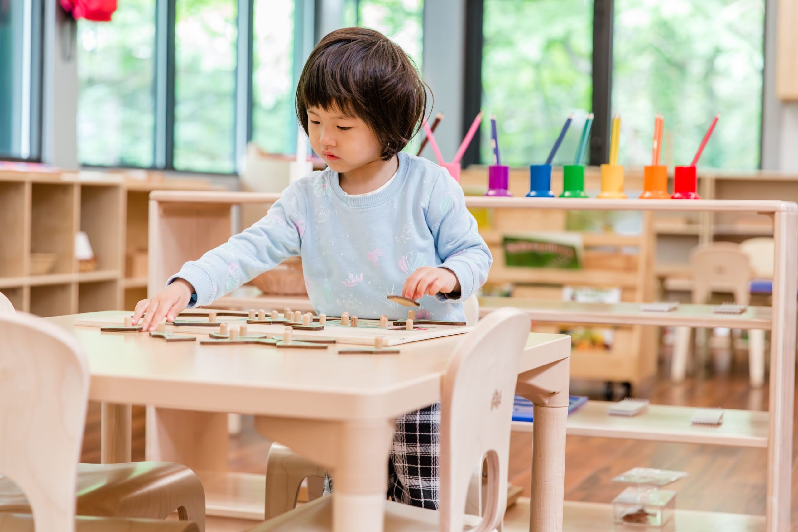 A girl in a blue shirt with black hair sits at a light wooden desk and moves light wooden puzzle pieces