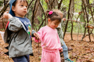 A young boy with a blue baseball cap and grey jacket walks through a wooded area with a stick in his hand. He is joined by a young girl in.a pink sweatsuit with pink bows on her ponytails.